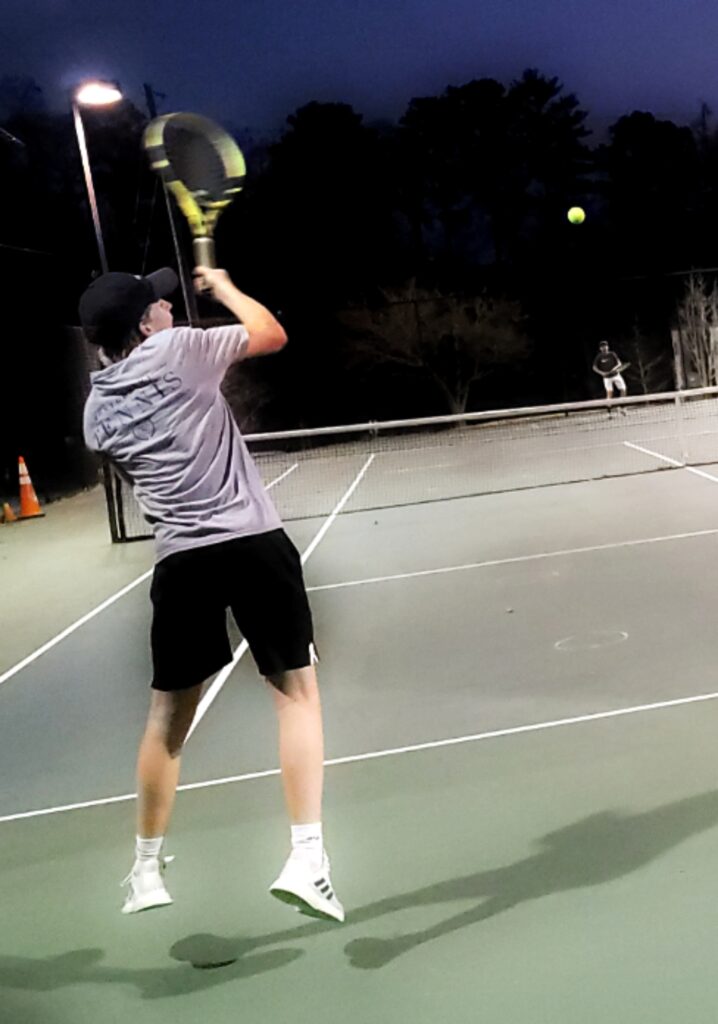 A tennis player in the foreground hangs in the air after returning the ball during a nighttime tournament.