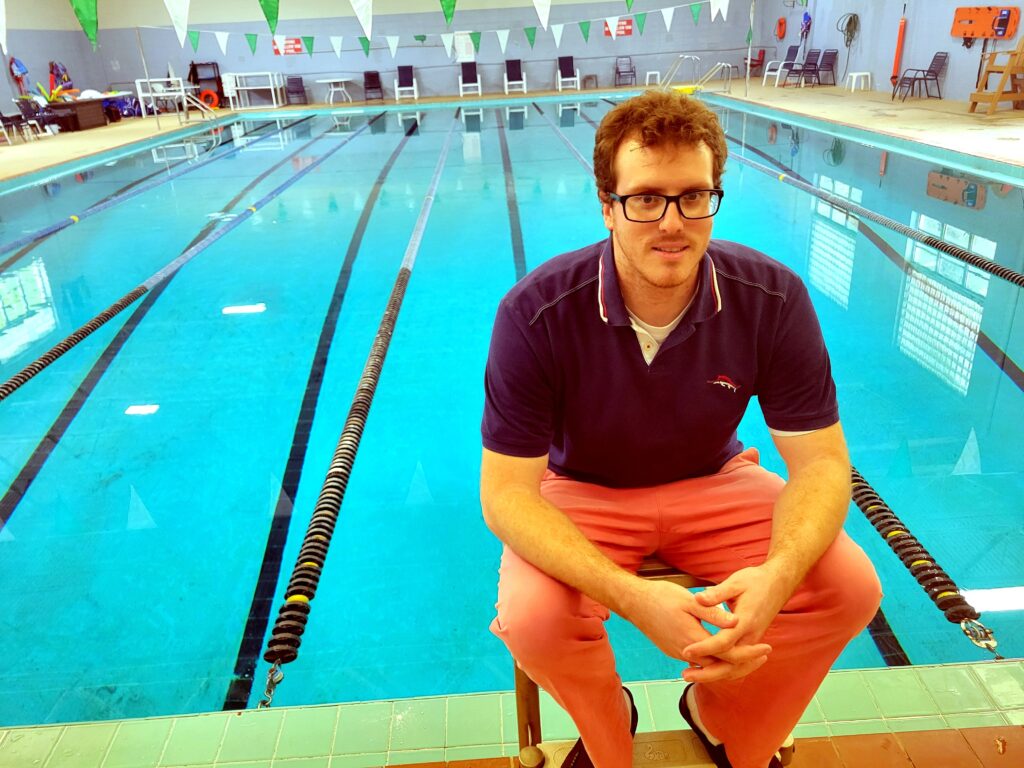 A man in a blue shirt, peach-colored pants, glasses, and red hair perches on one of the starting blocks with The J's indoor swimming pool filling the background