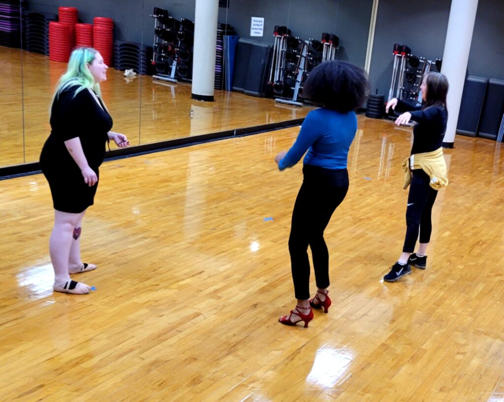 An instructor and two class participants practice ballroom dancing on a shiny wooden floor in front of a mirrored wall