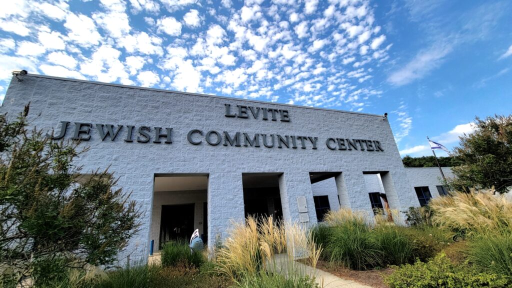 A white building that says "Levite Jewish Community Center," with brown and green vegetation in the foreground and blue sky with scattered clouds in the background