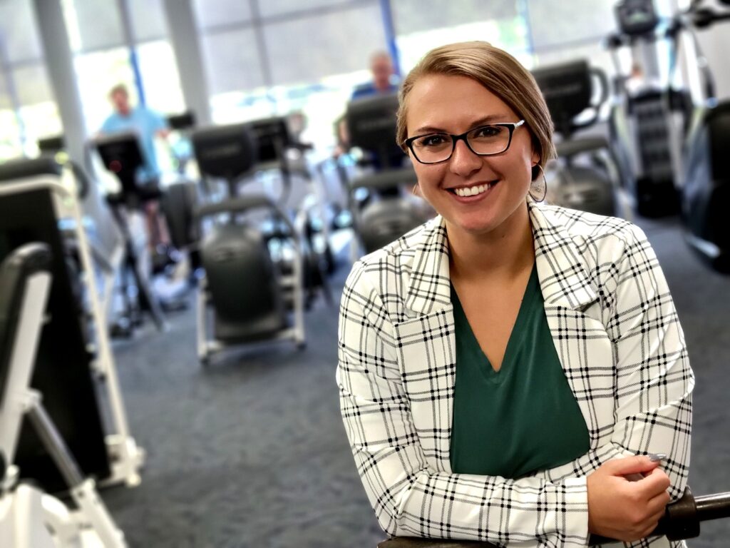 A woman in a checkered blazer and glasses poses on The J's Fitness Floor while members exercise on treadmills in the background