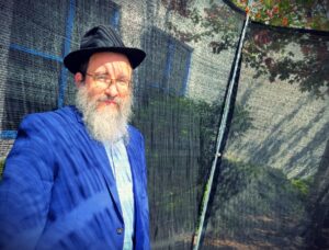 A bearded rabbi in a blue sportcoat and wearing a hat is shadowed by the roof of the sukkot he is standing under.