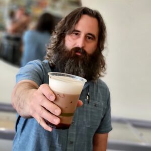 A bearded man extends a cup of freshly frothed egg cream drink toward the camera