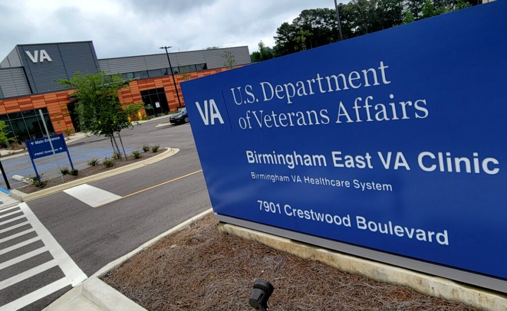 A photo of the blue Birmingham East VA Clinic sign with the VA building in the background