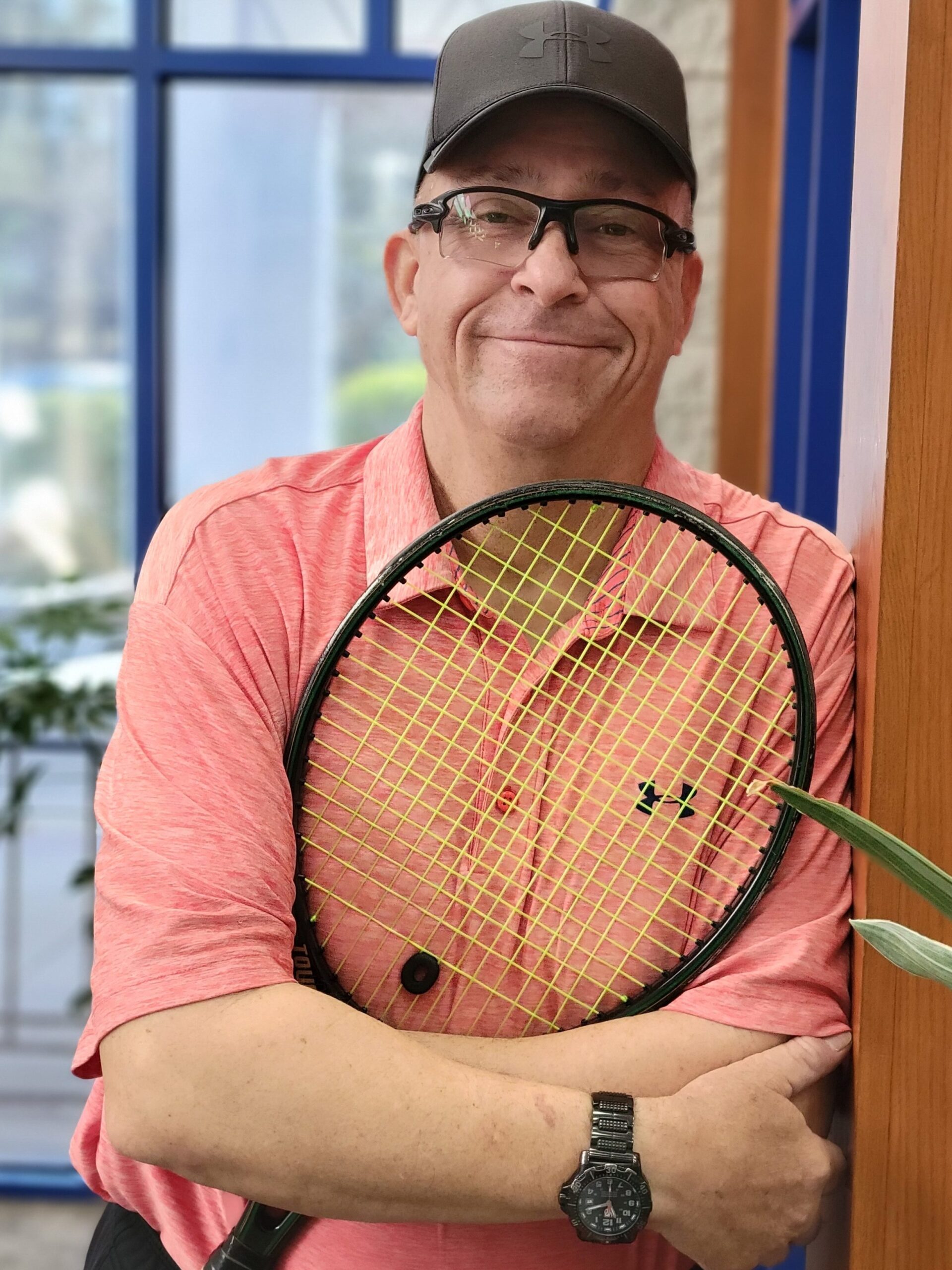 A man holding a tennis racket to his chest and wearing a cap leans against a wood-trimmed wall in a well-lit lobby