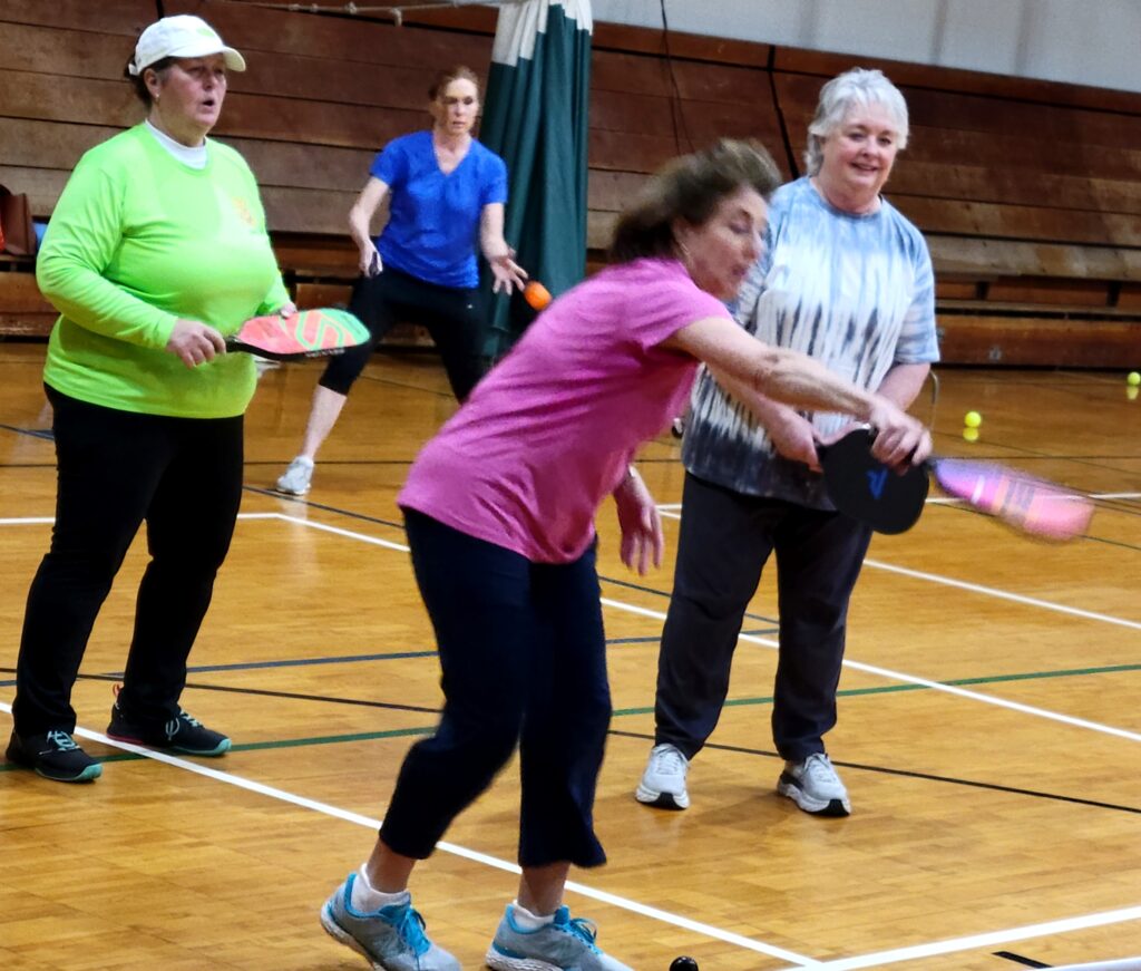 Four women on a court are practicing pickleball, with the woman closest to the camera, in a pink t-shirt, swinging her racket