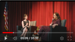 Two women are seated on a stage with a red-curtained background and an American flag draped on a pole to the left. One woman is interviewing the other.
