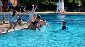 A group of seven kids in swimwear jump as a group from the edge of an outdoor swimming pool into the water